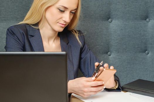 Beautiful woman holding a smartphone in hand while sitting at the wooden working table in home office.