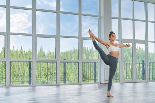 Active caucasian woman in sportswear balancing on one leg and stretching another during sport activity indoors. Spacious hall with panoramic windows.