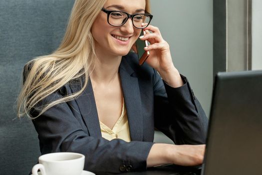 Beautiful young business woman with glasses is calling on mobile phone working on laptop in home office.