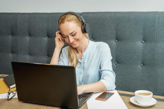 Happy young woman wearing headphones looking on laptop screen during online training with video conferencing at home office.