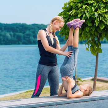 Little girt and woman are doing exercises on the grass at the shore of the lake.