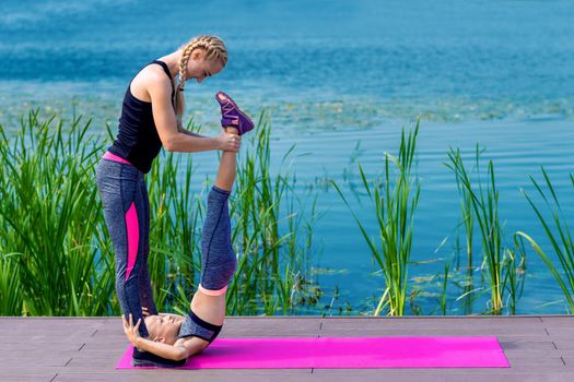 Little girt and woman are doing exercises on the grass at the shore of the lake.