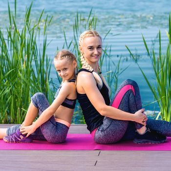 Portrait of mother and daughter sitting on yoga mat by the lake outdoors. Healthy and exercise concept.