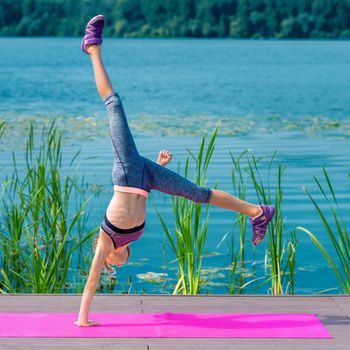 Young girl is doing a cartwheel outdoors on the roll mat by the lake.