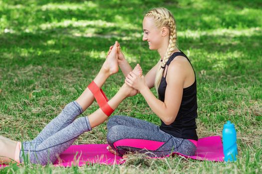 Young woman is training legs of little girl by fitness gum on mat in the park.