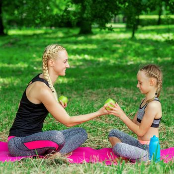Young woman and little girl in sportswear holding a green apples and sitting on the roll mat in park.