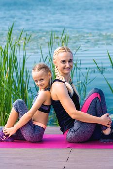 Portrait of mother and daughter sitting on yoga mat by the lake outdoors. Healthy and exercise concept.