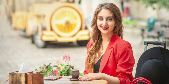 Outdoor portrait of young beautiful woman sitting at the table with cup of coffee and smartphone in the street.