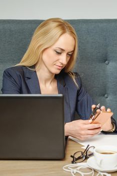 Busy attractive woman with blonde hair using smart phone working with laptop in office.