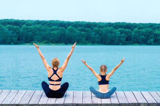 Mother and daughter doing yoga at the shore of the lake in summer.