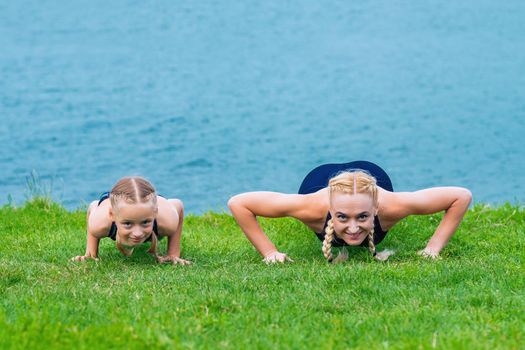 Little girt and woman are doing exercises on the grass at the shore of the lake.