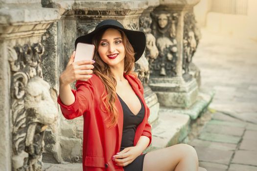 Happy young lady is taking photo of herself by smartphone on the city street.