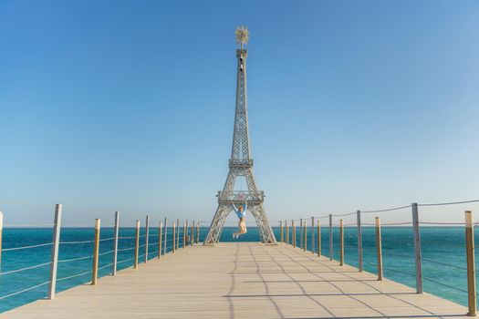 Large model of the Eiffel Tower on the beach. A woman walks along the pier towards the tower, wearing a blue jacket and white jeans