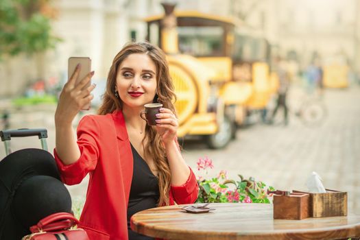 Beautiful brunette woman taking pictures of herself on a cellphone holding cup of coffee at cafe outdoors.