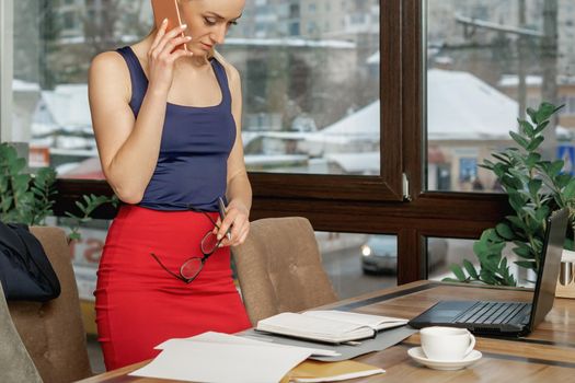 Young woman is talking by cell phone while working with documents in office.