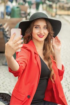 Happy young lady is taking photo of herself by smartphone on the city street.