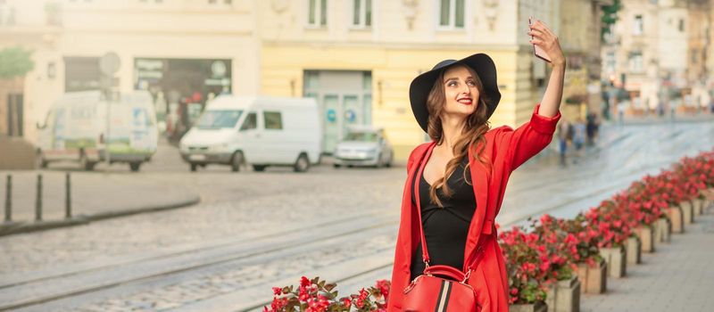 Stylish young woman is taking selfie walking on the street of old European city.