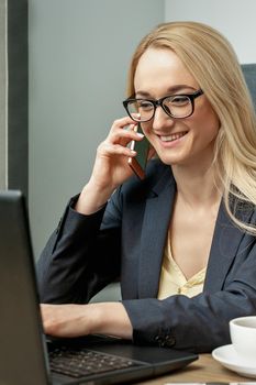 Young woman with glasses is talking by cell phone while working with laptop on sofa in home office.