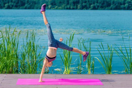 Cute girl is doing acrobatic handstand on roll mat by the lake.