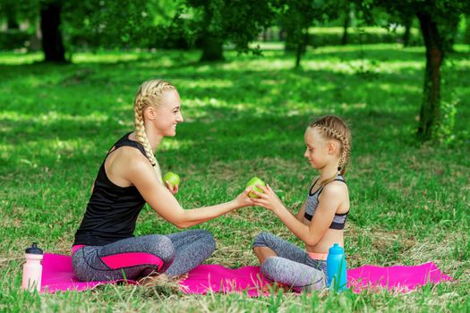 Mother and her daughter in sportswear holding a green apples and sitting on the roll mat in park.
