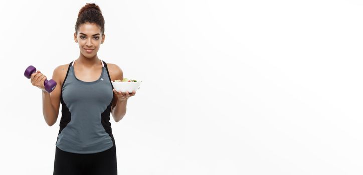 Healthy and Fitness concept - Beautiful sporty African American on diet holding dumbbell and fresh salad on hands. Isolated on white studio background