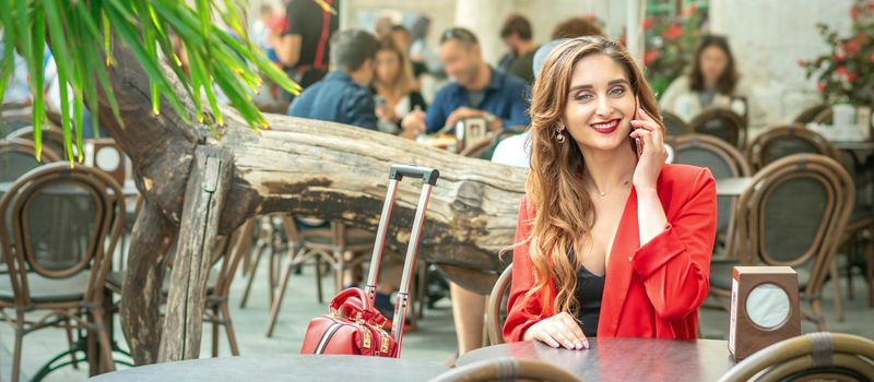 Young cute girl is talking by mobile phone sitting alone in coffee shop outdoors.