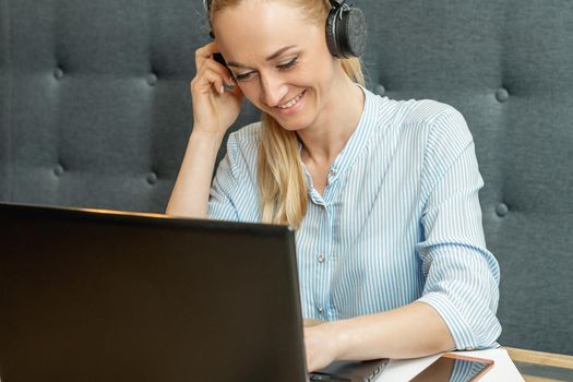 Happy young woman wearing headphones looking on laptop screen during online training with video conferencing at home office.