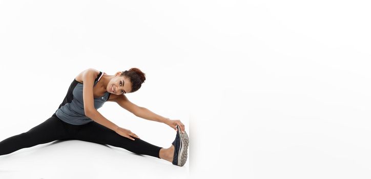 Sport, training, lifestyle and Fitness concept - portrait of beautiful happy African American woman stretching leg while sitting. Isolated on white studio background