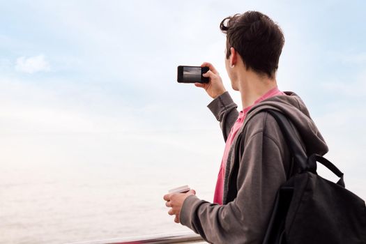 rear view of a young man with backpack taking a photo with a mobile phone, concept of technology and millennial lifestyle, copy space for text