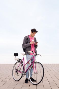 vertical photo of a young man with bicycle holding a coffee while using his mobile phone, concept of communication and sustainable urban transportation, copy space for text