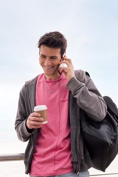 vertical photo of a happy young man with a coffee smiling while talking by mobile phone outdoors, concept of technology of communication and urban lifestyle, copy space for text