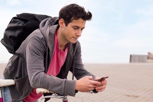 handsome young man with backpack leaning on a bicycle using a mobile phone, concept of communication and ecological urban transportation, copy space for text