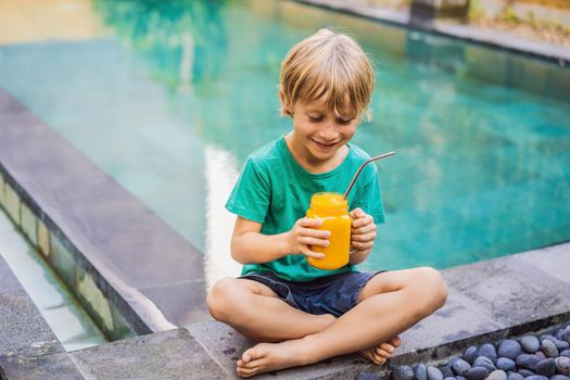 Boy drinking juicy smoothie from mango in glass mason jar with steel straw on the background of the pool. Healthy life concept, copy space.