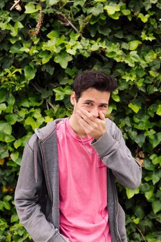 vertical photo of a nice young man laughing covering his mouth with one hand on a green plants background, concept of fun expressions and emotions, copy space for text