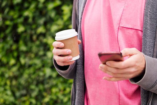 detail of the hands of an unrecognizable young man with a coffee using phone on a green plants background, concept of youth and technology of communication, copy space for text