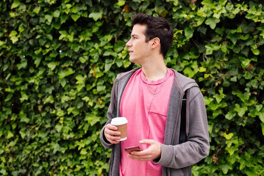 young teenager with a coffee using smart phone on a green plants background, concept of youth and technology of communication, copy space for text