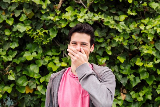 nice teenager laughing covering his mouth with one hand on a green plants background, concept of fun expressions and emotions, copy space for text