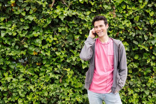 happy teen boy smiles while talking by phone on a green plant background, concept of youth and technology of communication, copy space for text