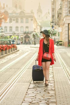 Beautiful young woman walking with a rolling suitcase on the tram track road in the old European city.