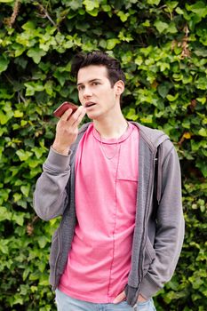 vertical photo of a young teen boy recording a voice message on mobile phone on a green plants background, concept of youth and technology of communication