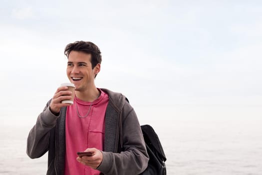 happy young man smiling with a coffee and using mobile phone outdoors, concept of technology and communication, copy space for text