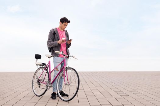 young man with bicycle holding a coffee while looking his mobile phone, concept of communication and sustainable urban transportation, copy space for text