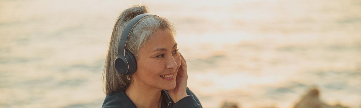Portrait of smiling woman in hoodie resting on the edge of seashore holding smartphone and listening to music on headphones