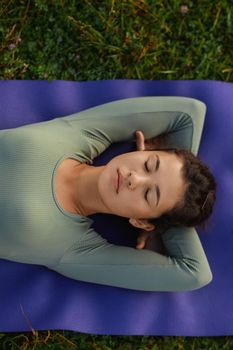 Crop of asian female woman closing eyes lying down on yoga mat among grass. Above view of relaxing girl in blue wear resting peacefully outdoors after doing yoga exercises. Concept of inner harmony.