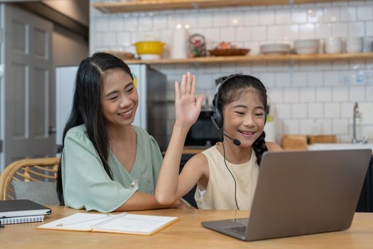 Happy asian mother and child sitting at kitchen table with colored pencils, attending virtual drawing class via video call, smiling and waving hello at laptop computer screen to greet online teacher.