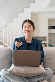 Beautiful asian woman sitting on couch in living room using laptop and watching something exciting interesting on television.