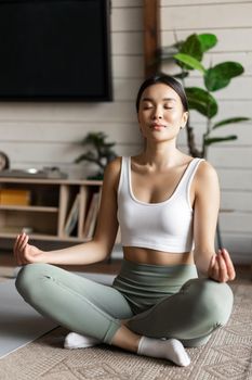 Mindfulness and meditation concept. Young asian woman doing workout at home, yoga meditation in living room on floor mat, getting focused.