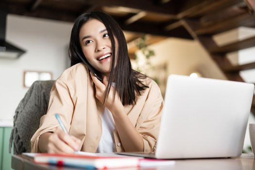 Young professional asian woman working from home, answer phone call, talking to client and writing down information, taking notes during conversation, sitting near laptop.