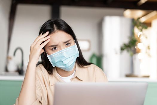 Sad asian girl in medical face mask working on laptop from quarantine, remote study during pandemic.
