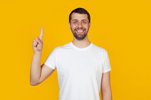 Modern young man with a beard in a white tank top shows number one with fingers on hand, smiling confidently and happily, looking at the camera. The man shows one finger. Number 1, yellow background